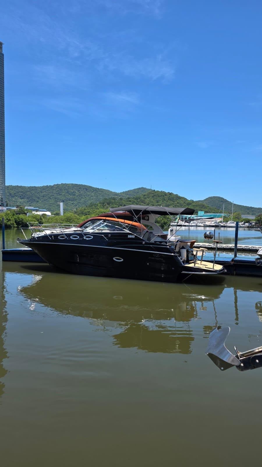 Lancha Regal 2660 preta atracada no píer da marina, com morros verdes e céu azul ao fundo